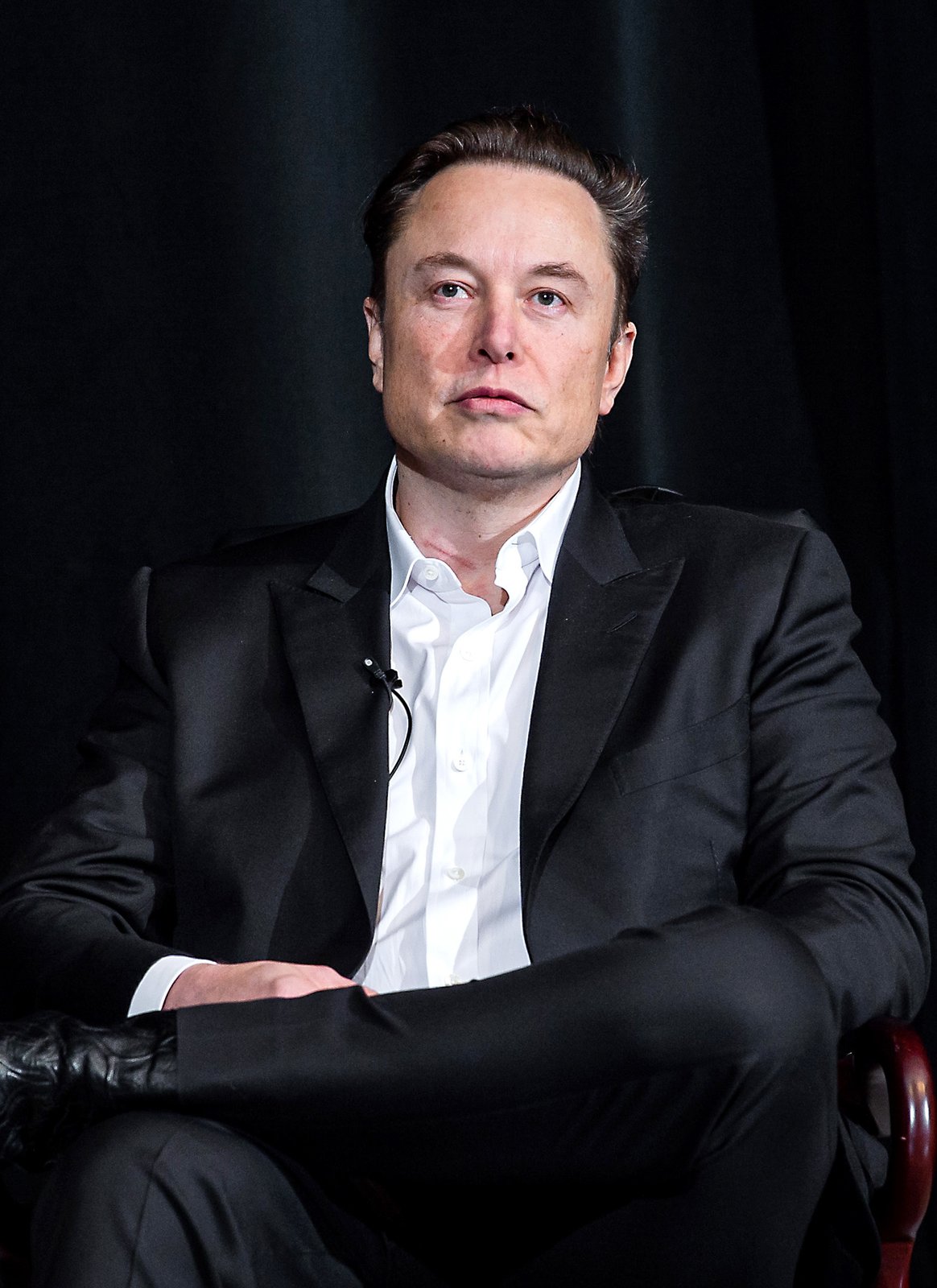 Elon Musk in 2022, seated against a dark curtain, listening to a question.